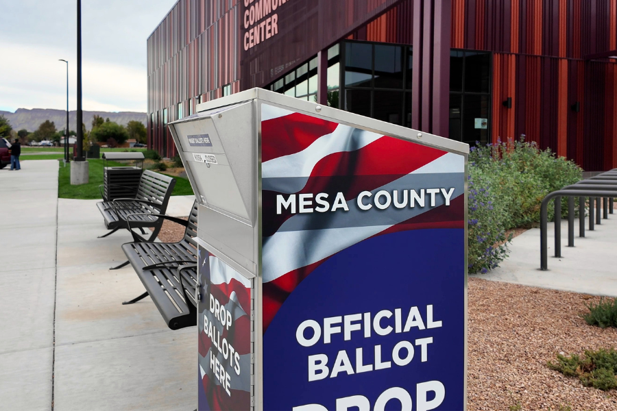 Photo of a metal ballot drop box with American flag graphics and the words “Mesa County” on the side. The red Clifton Community Center building is in the background.